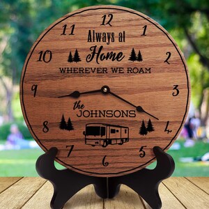 May include: A wooden clock with a black stand. The clock face has a wood grain pattern and features black numbers, a black silhouette of a camper, and the text "Always at Home Wherever We Roam" and "the Johnsons".
