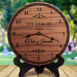 May include: A wooden desk clock with a black stand. The clock face is engraved with the words "The Desk Of" and a name. The clock hands are black and the numbers are in black.