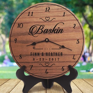 May include: A wooden clock with a brown wood grain finish. The clock face has black Roman numerals and a black inscription that reads "Baskin Finn & Heather 8~23~2017".