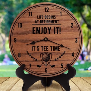 Puede incluir: Un reloj de madera con temática de golf. La esfera del reloj dice "La vida comienza en la jubilación, ¡Disfrútala! Es hora del tee". El reloj tiene un soporte negro y está colocado sobre un fondo borroso de hierba verde.