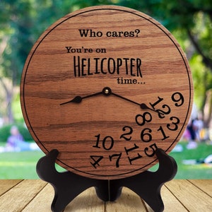 Op de afbeelding: Een houten klok met een zwarte standaard. De wijzerplaat van de klok heeft de tekst "Who cares? You're on HELICOPTER time..." en de cijfers 10, 2, 8, 4, 7, 11, 6, 1, 3, 9, 5 en 12.