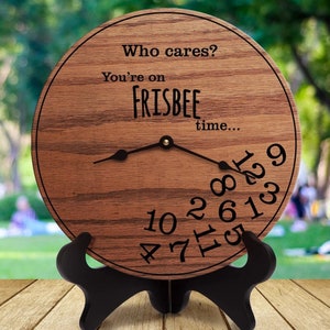 Puede incluir: Un reloj de madera con un acabado de grano de madera marrón. La esfera del reloj tiene el texto "Who cares? You're on FRISBEE time..." y los números del 1 al 12 están dispersos alrededor de la esfera. El reloj está en un soporte negro.