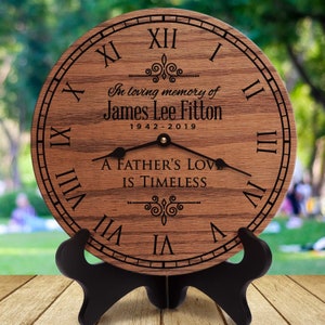 May include: A wooden clock with Roman numerals and a black stand. The clock face has the inscription "In loving memory of James Lee Fitton 1942-2019 A Father's Love Is Timeless".