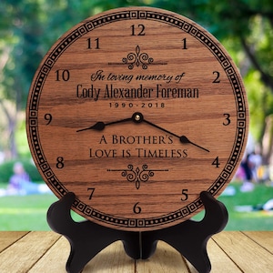 May include: A wooden clock with a black stand. The clock face is brown with black numbers and hands. The clock reads "In loving memory of Cody Alexander Foreman 1990-2018" and "A Brother's Love is Timeless".