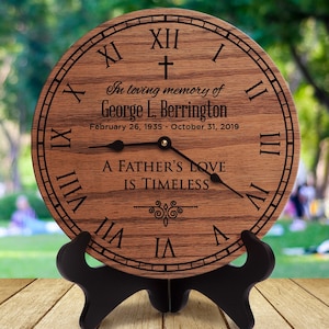 May include: A wooden clock with Roman numerals and a black stand. The clock face has the inscription "In loving memory of George L. Berrington February 26, 1935 - October 31, 2019 A Father's Love Is Timeless".