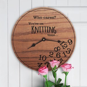 Puede incluir: Un reloj redondo de madera con el texto "Who cares? You're on knitting time..." y los números del 1 al 12 dispuestos en un círculo. Las manecillas del reloj apuntan a las 12 y las 3. El reloj está colgado en una pared blanca con rosas rosadas en primer plano.