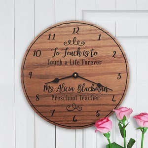 May include: A wooden clock with a brown wood grain finish. The clock face has black numbers and a quote that reads "To Teach is to Touch a Life Forever". The clock also has the name "Ms. Alicia Blackman" and the title "Preschool Teacher" written on it.
