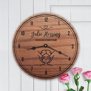 May include: A wooden clock with a brown wood grain finish. The clock face has black Roman numerals and a black outline. The center of the clock face has a black silhouette of a person receiving a facial massage. The text "Julie Herring Licensed Esthetician" is printed on the clock face.