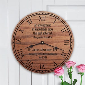 Puede incluir: Un reloj de madera con números romanos y una cita de Benjamin Franklin: "Una inversión en conocimiento paga el mejor interés." El reloj también incluye una inscripción personalizada: "Dr. James Alexander, DPT Universidad del Sur de California Abril 2019".