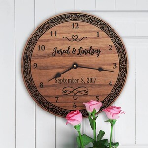 May include: A wooden clock with a black border and a decorative design. The clock face has black Roman numerals and the text "Jared & Lindsey" and "September 8, 2017".
