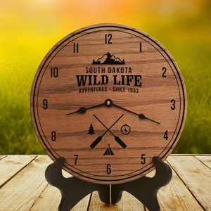 Peut inclure: Une horloge en bois avec un support noir. Le cadran de l'horloge présente un motif de chaîne de montagnes et le texte "South Dakota Wild Life Adventures - Since 1993". Le cadran de l'horloge comporte également une boussole, une tente et deux pagaies.