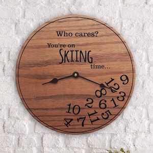 May include: Round wooden wall clock with black hands and numbers. The clock face has the text "Who cares? You're on SKIING time..." in black. The clock is made of wood with a visible grain pattern.