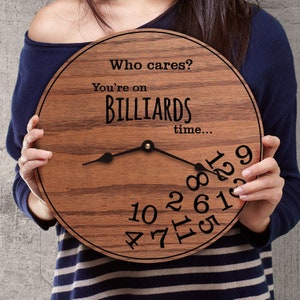 Könnte beinhalten: Eine runde Holzuhr mit braunem Finish. Das Zifferblatt der Uhr hat schwarze Zahlen und Zeiger, mit dem Text "Who cares? You're on BILLIARDS time..." auf dem Zifferblatt. Die Uhr wird von einer Person gehalten.