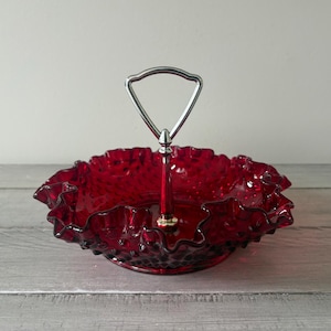 May include: A deep red glass serving dish featuring a hobnail texture and a ruffled, scalloped edge. The dish includes a central silver-toned metal handle with a triangular loop at the top, allowing for easy transport of the circular glass tray.