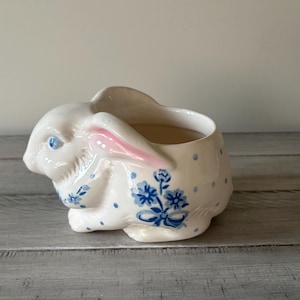 May include: A cream-colored ceramic planter shaped like a crouching rabbit. The rabbit features blue polka dots, a blue floral bouquet design on its side, pink-tinted inner ears, and bright blue eyes. The top of the rabbit has an open cavity.