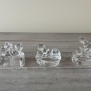 May include: Three clear glass figurines of cats in various poses. The cats are sitting on small, square or round bases. The glass is transparent, allowing light to pass through. The figurines are displayed on a gray wooden surface.