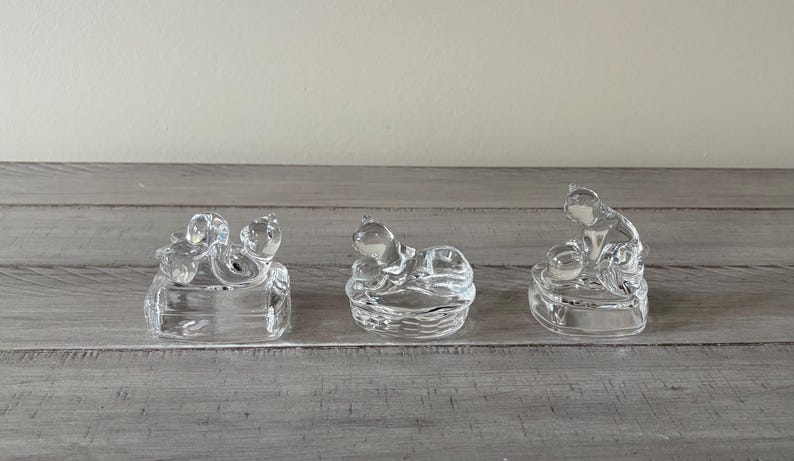 May include: Three clear glass figurines of cats in various poses. The cats are sitting on small, square or round bases. The glass is transparent, allowing light to pass through. The figurines are displayed on a gray wooden surface.