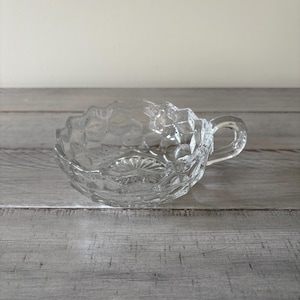 May include: A clear, decorative glass teacup with a handle. The cup has a textured, patterned design around the rim and on the base. The teacup is sitting on a gray wooden surface.
