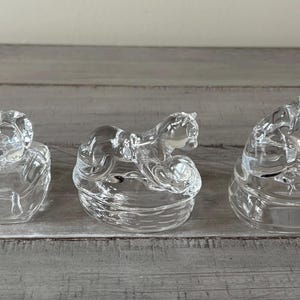 May include: Three clear glass figurines of kneeling figures. Each figure is kneeling on a rounded base. The figures are made of clear glass and are positioned on a gray wooden surface.