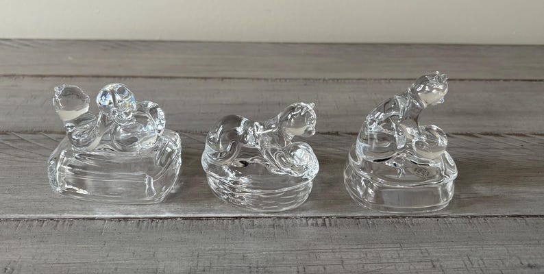 May include: Three clear glass figurines of kneeling figures. Each figure is kneeling on a rounded base. The figures are made of clear glass and are positioned on a gray wooden surface.