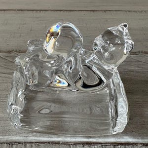 May include: A clear glass cat figurine with a rounded head and body, resting on a rectangular base. The glass reflects light, creating a rainbow effect. The cat's features are subtly defined, giving it a smooth, elegant appearance.