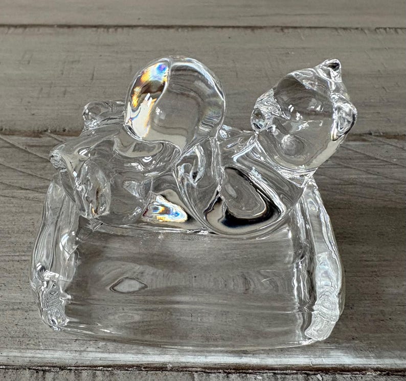 May include: A clear glass cat figurine with a rounded head and body, resting on a rectangular base. The glass reflects light, creating a rainbow effect. The cat's features are subtly defined, giving it a smooth, elegant appearance.