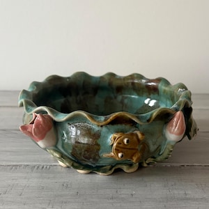 May include: A ceramic bowl with a ruffled edge, glazed in shades of teal and brown. The bowl is adorned with a frog and two pink and white lotus flower buds. The bowl sits on a light gray wooden surface.