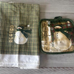 May include: A rectangular kitchen towel and a square potholder featuring a green and brown plaid pattern. Both items display a snowman illustration wearing a dark green scarf and the vertical text "Winter" in block letters on a light background.