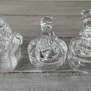 May include: Three clear glass frog figurines. Each frog is perched atop a round base. The frogs have detailed features, and the glass has a textured appearance. The figurines are displayed on a gray wooden surface.