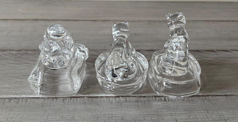 May include: Three clear glass frog figurines. Each frog is perched atop a round base. The frogs have detailed features, and the glass has a textured appearance. The figurines are displayed on a gray wooden surface.