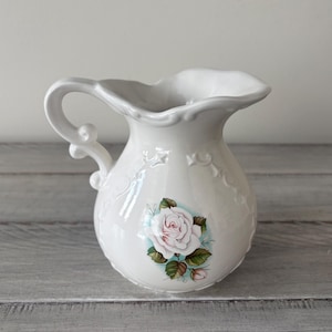 May include: White ceramic pitcher with a decorative handle and a floral rose design. The pitcher has a ruffled rim and embossed detailing. The rose is pink and green, adding a touch of color to the white pitcher.