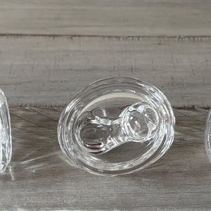 May include: Three clear glass candle holders with a decorative design. The candle holders are in different shapes: square, round, and oval. The candle holders are on a gray wooden surface.