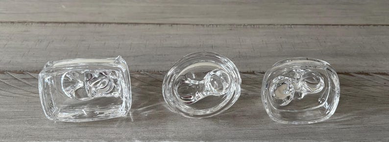 May include: Three clear glass candle holders with a decorative design. The candle holders are in different shapes: square, round, and oval. The candle holders are on a gray wooden surface.