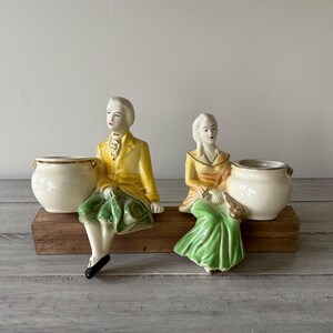 May include: Two ceramic figurines of seated figures, each holding a cream-colored pot. The figures are dressed in yellow and green attire, with the pots resting on a wooden base. The figurines have a vintage aesthetic.
