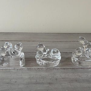 May include: Three clear glass figurines of cats in various poses. The cats are sitting on small, square or round bases. The glass is transparent, allowing light to pass through. The figurines are displayed on a gray wooden surface.