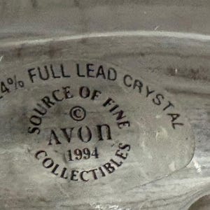 May include: A close-up of a clear, possibly crystal, object with a circular sticker. The sticker reads "OVER 24% FULL LEAD CRYSTAL SOURCE OF FINE AVON 1994 COLLECTIBLES."