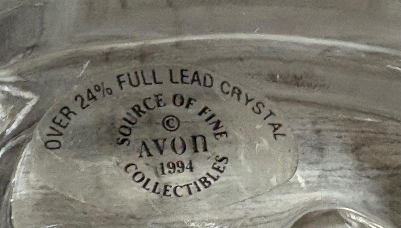 May include: A close-up of a clear, possibly crystal, object with a circular sticker. The sticker reads "OVER 24% FULL LEAD CRYSTAL SOURCE OF FINE AVON 1994 COLLECTIBLES."