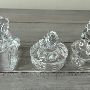May include: Three clear glass bear-shaped trinket boxes. Each box has a different bear pose. The boxes are made of clear glass and sit on a light gray wooden surface. The boxes are decorative.