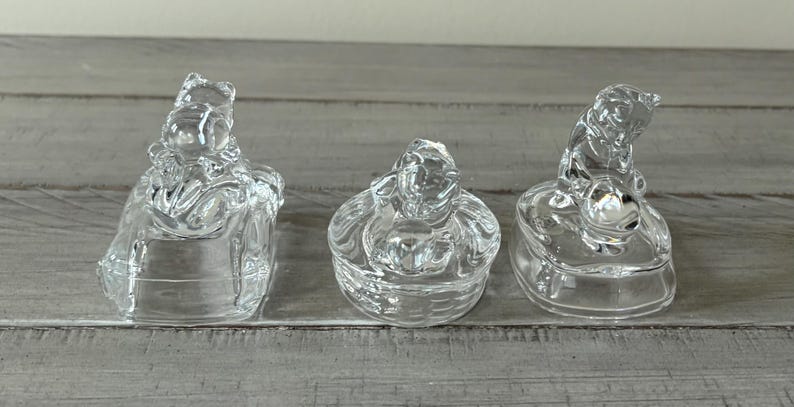 May include: Three clear glass bear-shaped trinket boxes. Each box has a different bear pose. The boxes are made of clear glass and sit on a light gray wooden surface. The boxes are decorative.
