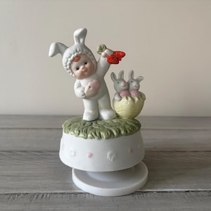 May include: A white ceramic Easter figurine featuring a child in a bunny suit holding a carrot and a heart. Two bunnies are in a yellow egg. The figurine sits on a white base with pink dots and floral designs.
