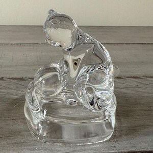 May include: A clear glass cat figurine with a seated pose. The cat is sculpted with smooth, flowing lines, and the glass reflects light, creating a sparkling effect. The figurine sits on a round base.