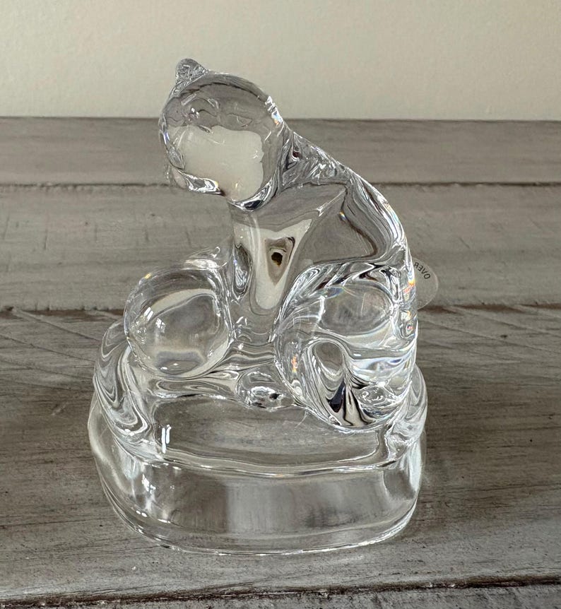 May include: A clear glass cat figurine with a seated pose. The cat is sculpted with smooth, flowing lines, and the glass reflects light, creating a sparkling effect. The figurine sits on a round base.
