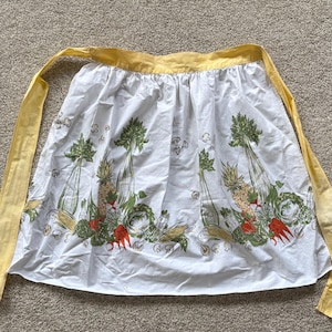 May include: A white waist apron featuring a yellow waistband and long ties. The fabric is decorated with a repeating print of green celery stalks, yellow pineapples, red peppers, corn, and small mushrooms arranged in a horizontal pattern.