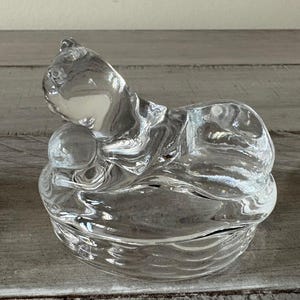 May include: A clear glass cat figurine with a curled-up body and a small ball. The cat is resting on a round, textured base. The glass has a rippled effect, creating a unique visual texture. The figurine is sitting on a wooden surface.
