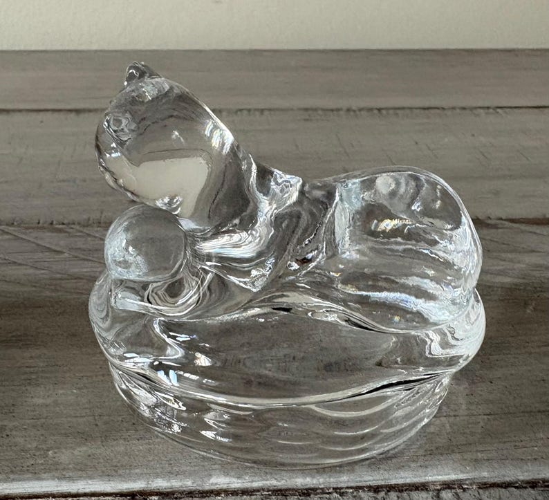 May include: A clear glass cat figurine with a curled-up body and a small ball. The cat is resting on a round, textured base. The glass has a rippled effect, creating a unique visual texture. The figurine is sitting on a wooden surface.