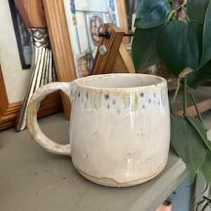 May include: A handmade ceramic mug with a rounded body and a curved handle. The mug has a speckled, off-white glaze with blue and green accents near the rim. The mug is sitting on a gray surface.