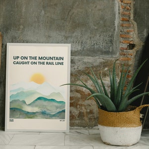May include: A white framed print with a watercolor illustration of a mountain range with a yellow sun. The text "UP ON THE MOUNTAIN CAUGHT ON THE RAIL LINE" is printed in blue and green.