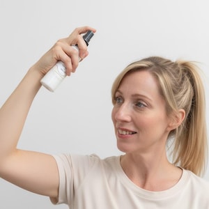 May include: A white spray bottle with a black nozzle is held up in the air. The bottle has a label with text. The person in the image is wearing a light beige t-shirt.