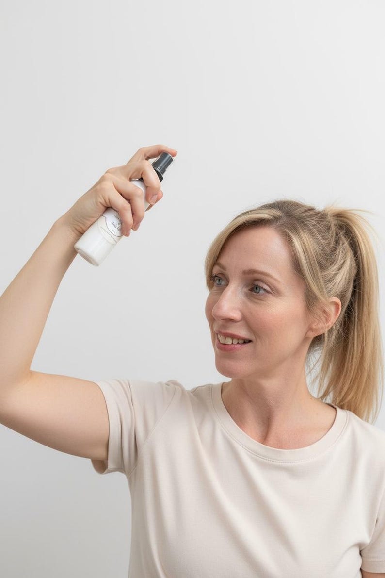 May include: A white spray bottle with a black nozzle is held up in the air. The bottle has a label with text. The person in the image is wearing a light beige t-shirt.
