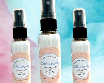 May include: Three clear spray bottles filled with a white liquid. Each bottle has a black cap and a clear spray nozzle. A peach-colored label with the words "Cotton Candy Hair Fragrance & Body Mist" is on each bottle. The background is a mix of blue and white.
